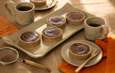 Foto em tons de marrom da receita de torta de ganache de cacau servida em diversas porções sobre uma base de cerâmica retangular e uma sobre um pires de cerâmica, além de duas caneca de mesmo material ao lado