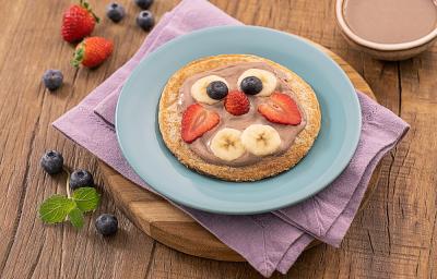 Foto da receita de panquequinha de cenoura em formato de "rostinho" com as frutas, servida em um prato de porcelana azul claro sobre um paninho lilás em uma tábua de madeira. Ao fundo algumas frutas espalhadas e um bowl de porcelana com cacau em pó.