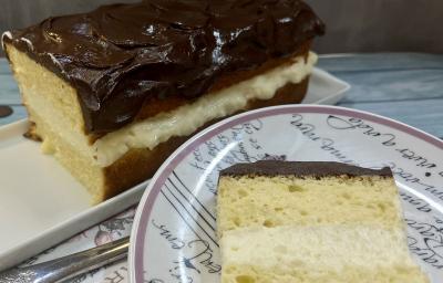 Foto da receita de Boston Pie. Observa-se o bolo retangular com uma camada de creme branco no meio e cobertura de chocolate no plano de fundo. À frente, um prato de sobremesa redondo com uma fatia do bolo.