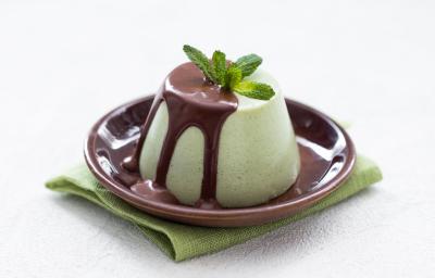 Fotografia de uma mousse de menta com calda de chocolate, com folhas de menta por cima. A mousse está em um prato pequeno na cor marrom, sobre um pano dobrado, na cor verde, que está sobre uma mesa branca.