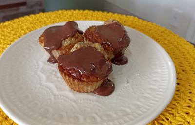 Fotografia em tons de amarelo em uma bancada de pedra, um apoia prato amarelo de renda, um prato branco com três bolinhos de banana com calda de chocolate em cima.