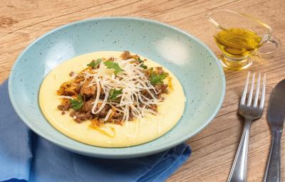 Fotografia de um prato fundo azul claro com uma polenta cremosa, e por cima, uma carne moída com cheiro verde e queijo parmesão ralado. O prato está apoiado em um pano azul pequeno que está sobre uma mesa de madeira, e ao lado tem talheres e azeite.