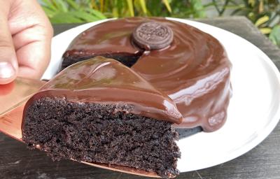 Foto aproximada de um bolo de chocolate com a fatia na frente sobre uma faca. O bolo está decorado com uma calda de chocolate e com um biscoito Negresco no centro.