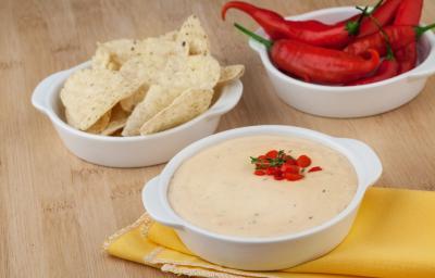 Fotografia em tons de amarelo em uma bancada de madeira clara, um paninho amarelo, um potinho branco com o patê de batata, outro potinho branco com tortillas e ao fundo um potinho com pimenta de dedo-de-moça.