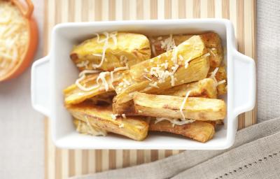 Fotografia em tons de bege em uma mesa de madeira com uma toalha bege e uma travessa retangular branca com as mandiocas assadas com parmesão dentro.