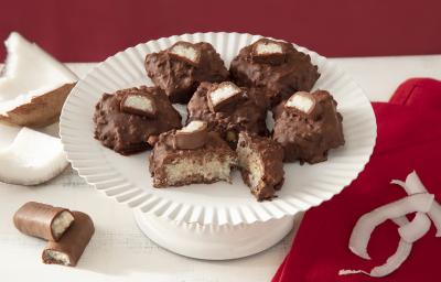 Foto da receita de cocada de prestígio. Observa-se uma boleira branca com 6 cocadas banhadas no chocolate. A da frente está cortada e é possível ver o coco de dentro