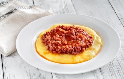 fotografia em tons de branco, amarelo e vermelho de uma bancada branca vista de cima. Contém um prato redondo branco com polenta, e por cima um molho bolonhesa.