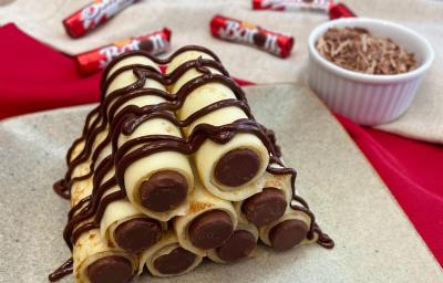 Fotografia em tons de vermelho com uma torre de crepes recheados ao centro. Os crepes encontram-se empilhados e recheados com chocolate Baton ao Leite e cobertos com uma calda de chocolate.