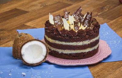 Foto da receita de Bolo Naked de Prestígio, decorado com pedaços de chocolate, sobre um prato rosa, numa bancada de madeira com tecidos azuis e dois pedaços de coco fresco aberto