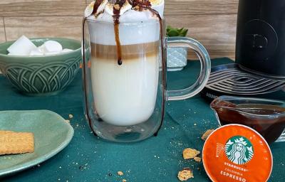 Fotografia em tons de verde em uma bancada com um pano verde, uma xícara de vidro grande com a bebida de leite e café preparada dentro dela. Decorada com marshmallow, calda de chocolate e biscoito triturado.