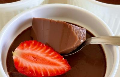 Fotografia de uma receita de gelatina de chocolate dentro de um recipiente branco fundo e médio. Tem uma colher segurando uma poção da receita e ao lado, um pedaço de morango. Fotografia de uma receita de gelatina de chocolate dentro de um recipiente branco fundo e médio. Tem uma colher segurando uma poção da receita e ao lado, um pedaço de morango.