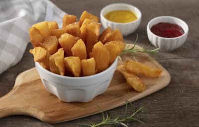 Fotografia em tons de amarelo em uma bancada de madeira escura, uma tábua de madeira, um pote branco fundo com várias mandiocas fritas. Ao fundo, um pano bege xadrez e dois potinhos um com ketchup e um de mostarda.