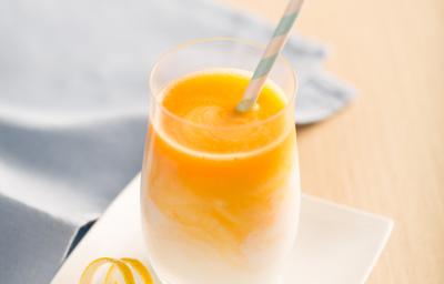 Fotografia em tons de laranja e azul em uma bancada de madeira clara, um pano azul, um prato pequeno branco quadrado com um copo de vidro em cima dele e o smoothie de laranja, manga e cenoura dentro dele.