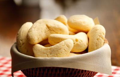 Fotografia em tons de vermelho em uma bancada de madeira, um pano vermelho xadrez, uma cesta de vime marrom, um guardanapo branco de pano com as chipas, pães à base de ovos, manteiga e queijo, dentro dela.