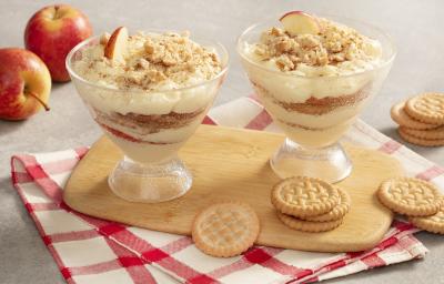 Fotografia em tons de cinza, vermelho e branco de uma bancada cinza com um paninho xadrez branco e vermelho com uma tábua de madeira, sobre ela duas taças com pavê de maçã e biscoitos tostines espalhados pela mesa. Ao fundo duas maçãs.