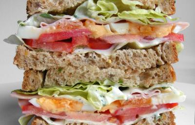 Fotografia de um sanduíche em camadas no pão de forma integral com tomate, repolho, maionese, presuntada e alface. O sanduíche está cortado na metade, sendo uma em cima da outra, que está sobre uma mesa branca.