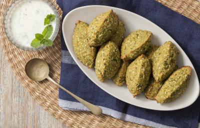 Fotografia em tons de verde, azul e branco, ao centro uma travessa branca com os bolinhos de falafel, ao lado para acompanhar um potinho de vidro com o molho de iogurte e uma folhinhas verdes para decorar. Abaixo está um guardanapo azul.