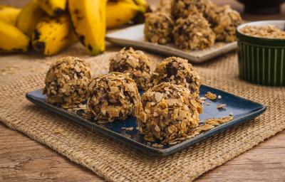 Fotografia em tons de amarelo e azul em uma bancada de madeira, um jogo americano de palha, um recipiente retangular azul com alguns brigadeiros de banana coberto com cereais Neston. Ao fundo, um cacho de banana.