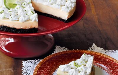 Fotografia em tons de vermelho e branco, com boleira vermelha contendo uma torta com creme e cobertura brancos, ao lado prato branco com borda vermelha com um pedaço de torta sob uma espátula e sobre guardanapo branco, tudo sobre bancada de madeira.