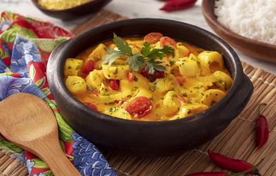 Fotografia em tons de amarelo em uma bancada de madeira de cor branca. Ao centro, uma panela preta contendo a moqueca. Ao lado, há um pano florido vermelho com uma colher de pau em cima. Ao fundo, há uma tigela de arroz.