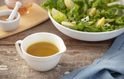 Fotografia em tons de azul em uma bancada de madeira escura, um pano azul, uma tábua de madeira, um potinho branco com o molho de mostarda e mel dentro. Ao fundo, um recipiente com salada verde para acompanhar o molho e uma tábua com mais potinhos.