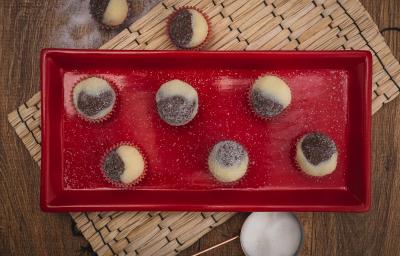 Fotografia em tons de vermelho em uma bancada de madeira escura, um jogo americano de palha, um recipiente retangular vermelho com brigadeiros mesclados de brigadeiro branco e preto em forminhas de vermelho.