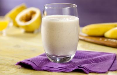 Fotografia em tons de roxo, pardo, branco e amarelo de uma bancada vista de frente. Contém um pano roxo com um copo transparente por cima com a vitamina. Ao junto maracujá cortado e bananas