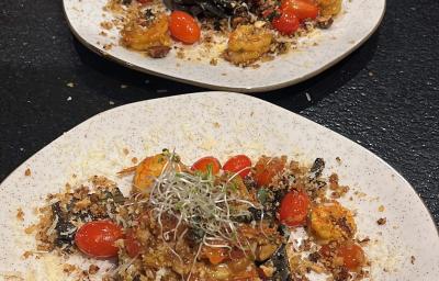 Fotografia de dois pratos de vidro parcialmente quadrados, de pontas arredondadas, sobre uma mesa preta. Dentro dos pratos tem uma massa linguine nero com molho rústico de tomate grape, camarões e farofa de castanhas.