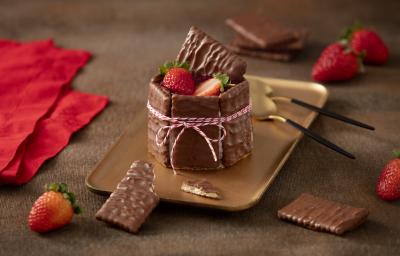 Foto de uma bancada escura com alguns tecidos vermelhos, morangos e biscoitos cobertos de chocolate espalhados. Ao centro há um prato retangular dourado com a receita pronta montada e decorada, acompanhada de duas colheres douradas e pretas