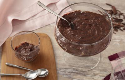 Foto da receita de Mousse Fácil de Chocolate. Observa-se um recipiente redondo com a mousse e, ao lado, um recipiente individual. Também tem duas colherinhas ao lado e raspas de chocolate como decoração.