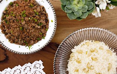 fotografia em tons de marrom e branco tirada de uma bancada marrom vista de cima. Contém um recipiente ao lado esquerdo redondo branco com a farofa e ao lado direito um recipiente redondo transparente com arroz