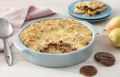 Fotografia em tons de azul em uma bancada de madeira clara, um paninho bege, um recipiente azul claro com o crumble de pera com calipso. Ao fundo, um prato com um pedaço do crumble, peras e calipso espalhados pela mesa.