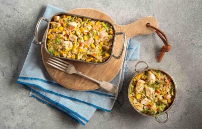 Foto em tons de cinza, azul e marrom uma bancada cinza vista de cima, um pano azul claro, uma tábua de madeira redonda com um recipiente retangular com alças que contém arroz com pedaços de frango, um garfo e um recipiente redondo com arroz e frango