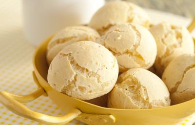 Fotografia em tons de amarelo em uma bancada de madeira esverdeada, um paninho branco com bolinhas amarelas, um recipiente amarelo com vários pães de queijo dentro dele. Ao fundo, uma xícara branca com café.