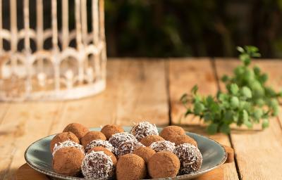 Fotografia em tons de verde em uma bancada de madeira com um prato raso verde ao centro apoiado em uma tábua de madeira e os brigadeiros em cima. Ao lado, brigadeiros espalhados pela bancada.