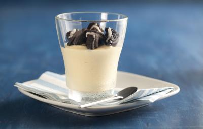 fotografia em tons de azul e branco de uma bancada azul vista de frente, contém um pratinho branco com um pano em tons de azul branco, um copo transparente com creme de maracujá com pedaços de biscoitos por cima.