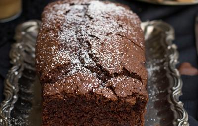 Fotografia em tons de marrom de uma bancada marrom vista de cima, contém uma bandeja de alumínio com um bolo inglês de chocolate com açúcar polvilhado por cima.