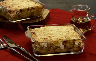 Foto em tons de vermelho da receita de delícia de bacalhau servida em duas porções em refratários de vidros um atrás do outro. Ao lado há um garfo e uma faca prateados e ao fundo uma jarra de vidro com azeite, tudo sobre uma mesa vermelha