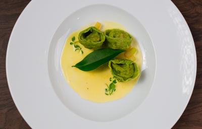 Fotografia em tons de branco com um prato branco ao centro. Em cima do prato existe uma receita de massa recheada de cor verde acompanhada de um molho de cor amarela.