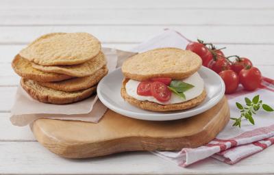 Fotografia em tons de branco, marrom e vermelho, ao centro fatias de pão sobre guardanapo e prato branco com lanche contendo queijo branco e tomate sobre tábua, ao lado guardanapo branco com listras vermelhas e tomates-cereja, tudo sobre bancada branca.