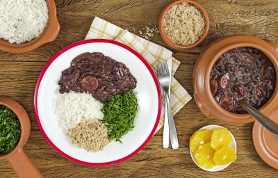 Fotografia em tons de marrom e vermelho de uma bancada de madeira, sobre ela recipientes redondos com feijoada, arroz, couve e farofa. Ao centro um paninho branco xadrez com um prato branco redondo com um prato feito de feijoada.