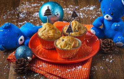 Fotografia em tons de laranja e azul em uma bancada de madeira, um prato laranja com três muffins em cima dele. Ao lado, enfeites e decoração natalina.