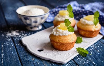 Fotografia de três muffins de abacaxi com decoração de um creme branco, abacaxi e folhas de hortelã sobre uma mini tábua na cor branca. Ao fundo, o creme branco dentro de um recipiente fundo nas cores azul e branco, sobre uma mesa de madeira na cor azul.