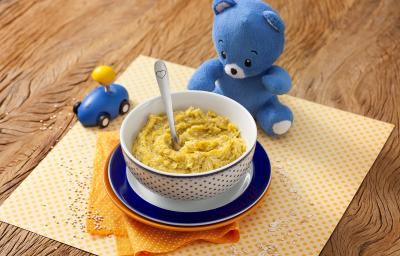 Fotografia em tons de laranja em uma bancada de madeira com um paninho amarelo de bolinha no centro e um potinho branco com bolinhas azuis com a sopinha de frango com abóbora. Ao lado, um ursinho de pelúcia azul e um carrinho azul de brinquedo.