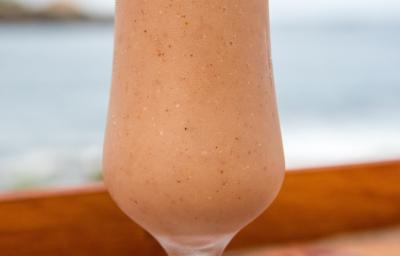 Fotografia de um copo de vidro, estilo taça, com uma bebida de suco de goiaba e neston sobre uma mesa de madeira com vista para a praia.