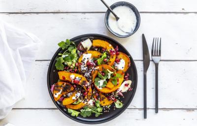 Fotografia vista de cima de uma salada de folhas com molho de laranja, pedaços de abóbora, sementes e um ramo de coentro em um prato raso preto. Ao lado tem talheres e um molho de iogurte em um pote pequeno de vidro azul, com uma colher apoiada.