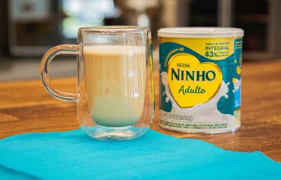 Fotografia em tons de azul com uma caneca de virdro ao centro. Dentro da caneca existe um cappuccino de cor caramelo e ao lado existe uma lata de leite NINHO Adulto em pó