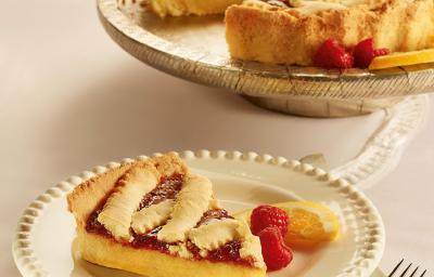 Fotografia em tons de bege em uma mesa de madeira, um guardanapo de pano bege e branco, um prato redondo branco com detalhes bege e uma fatia da crostata com recheio de laranja. Ao fundo, um prato redondo maior com a crostata inteira.