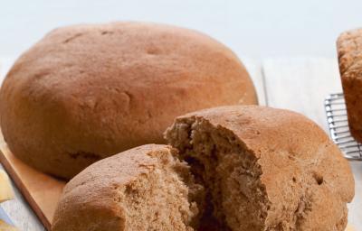 Fotografia em tons de marrom em uma bancada de madeira com uma tábua de madeira clara e o pão preto redondo grane apoiado nela, e um outro pedaço de pão já cortado.