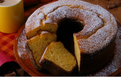 Fotografia em tons de vermelho em uma bancada de madeira escura com um pano laranja e vermelho xadrez, um suporte para bolo com o bolo de milho em cima dele cortado em algumas fatias. Ao lado, xícaras coloridas com cappuccino dentro.